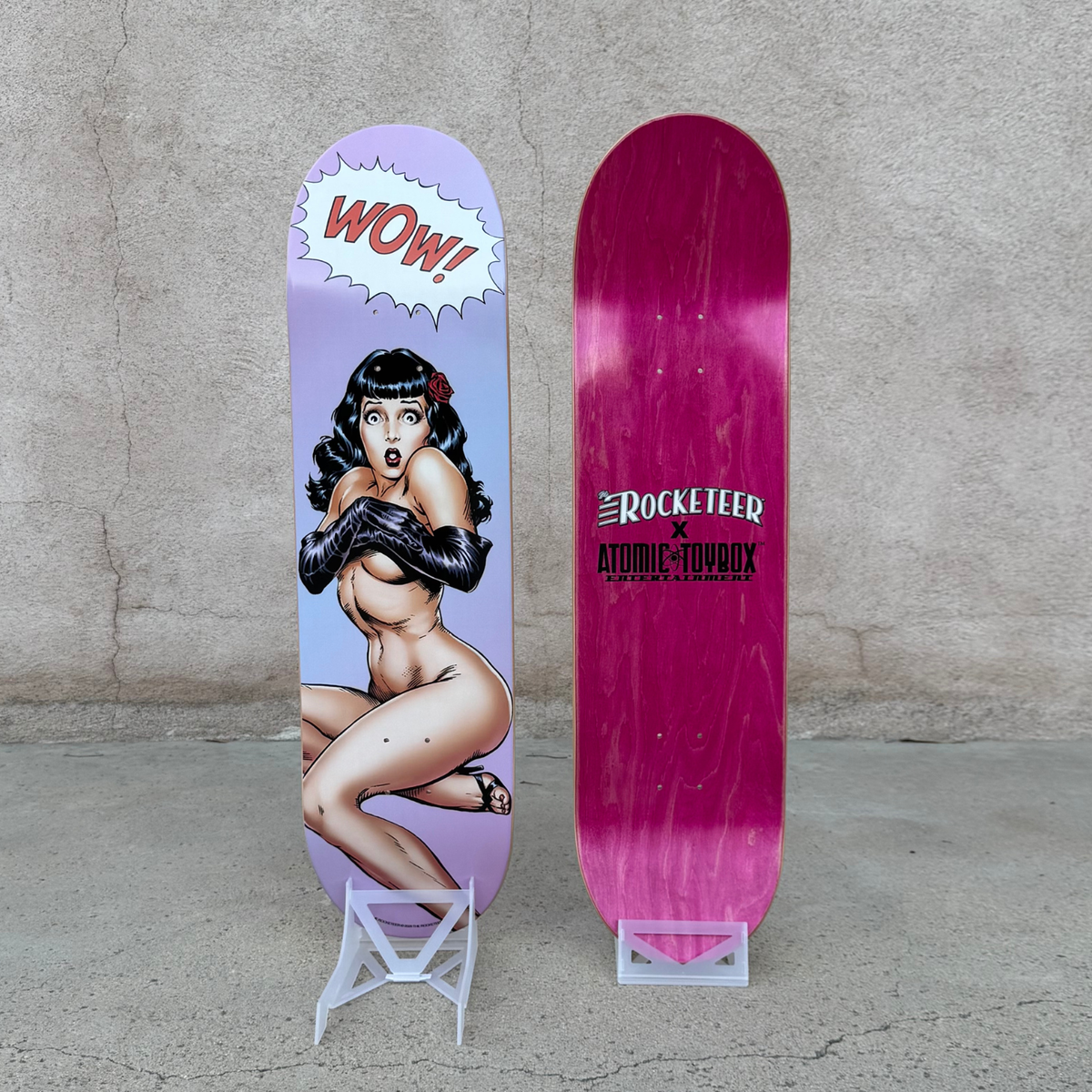 Two skateboard decks displayed upright on small white stands against a concrete wall. The left deck has a lavender background with a pin-up style illustration of a nude woman and a comic-style 'WOW!' speech bubble.