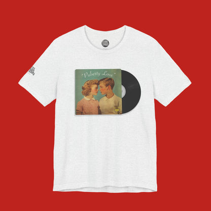 A white heather crewneck t-shirt featuring a graphic print of a vintage CD album cover titled 'Puberty Love' with a black vinyl record overlay, displayed on a clean white background.