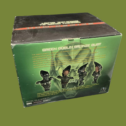 A boxed collectible figure package labeled 'Green Goblin Bronze Bust' sitting on a glass surface. The box is predominantly green with black top panels, product text, and illustrations of the Green Goblin busts.