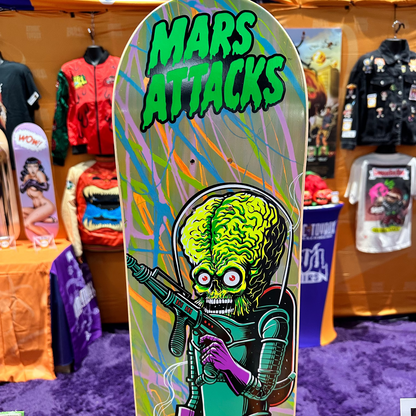 A skateboard deck featuring a colorful retro sci-fi alien graphic with the text 'MARS ATTACKS' in green, displayed upright at a convention booth with merchandise in the background.