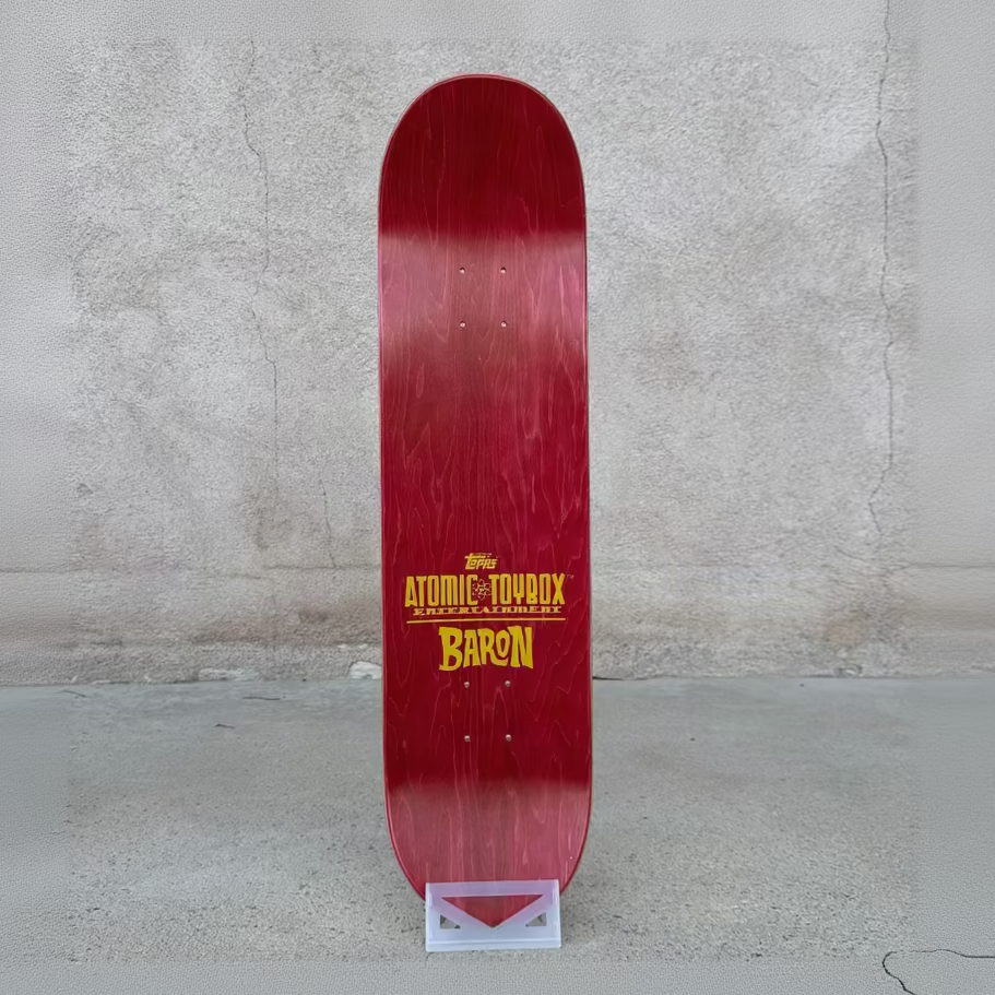 A red skateboard deck standing vertically on a small display stand against a textured concrete wall and floor. The deck features yellow and white text reading 'Atomic Toxix' and 'Baron'.