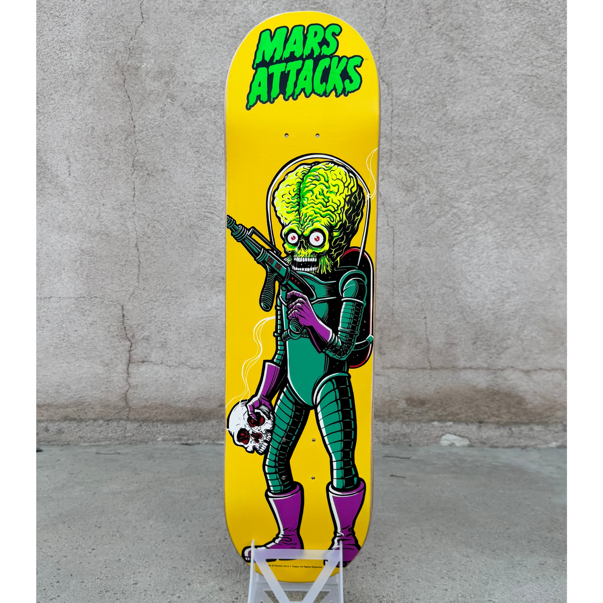 A skateboard deck with a bright yellow background and a green alien graphic holding a skull and gun, with the text 'MARS ATTACKS' in green at the top. The deck is displayed upright on a small white stand against a gray concrete wall and floor.