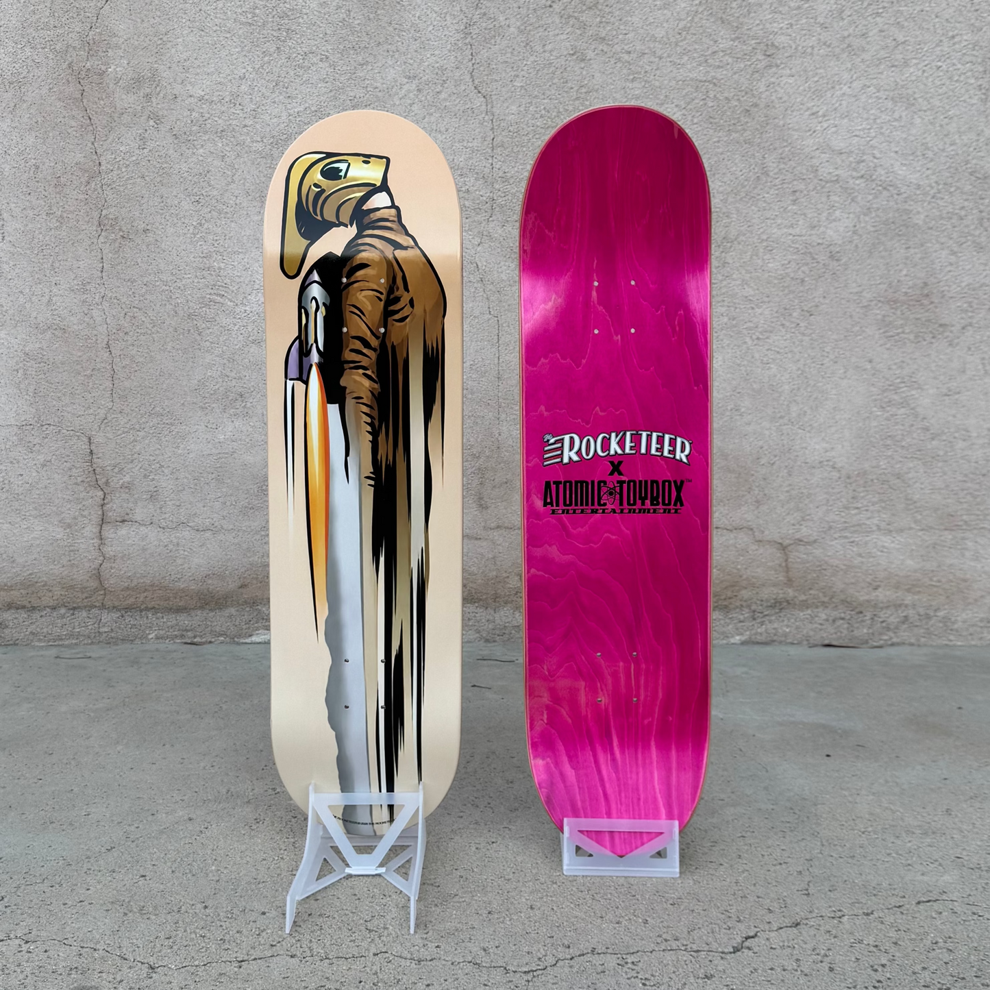 Two skateboard decks displayed upright on small stands against a concrete wall and floor. One deck has a cream base with a graphic of a bird of prey in a suit, and the other is a bright hot pink deck with white and black text reading Atomic Toybox and The Rocketeer